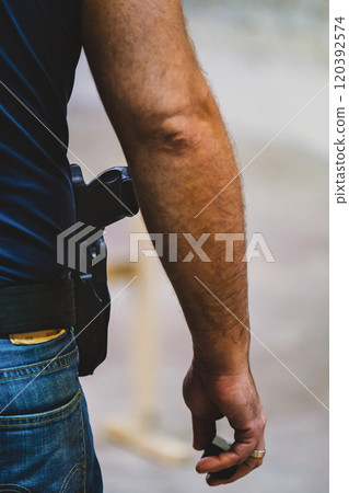 close-up of a person's arm and hip area.Man is wearing jeans and has a holster with a firearm attached to their belt. The hand appears to be holding an object, but it's not clearly visible  120392574