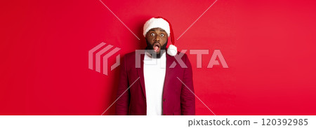 Christmas, party and holidays concept. Shocked Black man in santa hat drop jaw and stare at camera, stand against red background 120392985