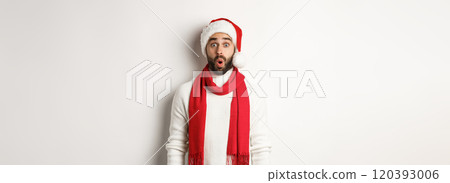 Christmas holidays. Bearded man looking surprised at camera, wearing santa party hat and red scarf, standing against white background 120393006