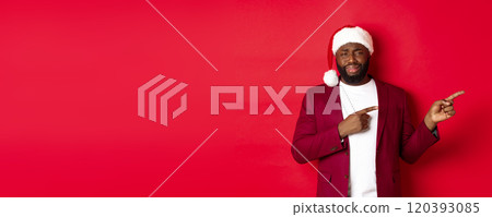 Christmas, party and holidays concept. Skeptical and unamused Black man looking with disdain, pointing fingers right at logo, standing in santa hat against red background 120393085
