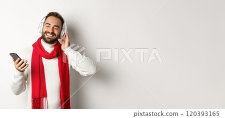 Winter holidays and technology concept. Satisfied man listening music in headphones with closed eyes, smiling with pleasure, holding smartphone, white background 120393165