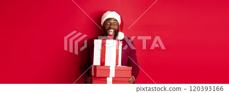 Christmas, New Year and shopping concept. Happy Black man in santa hat and blazer holding xmas presents, bring gifts and smiling, standing against red background 120393166