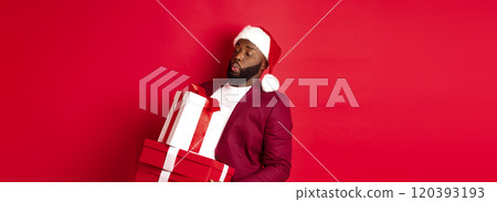 Christmas, New Year and shopping concept. Funny african american man in santa hat carry heavy xmas presents, holding gifts, standing over red background 120393193