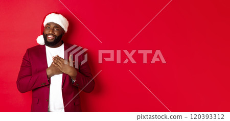 Christmas, party and holidays concept. Grateful african american man in santa hat saying thank you, holding hands on heart and smiling, feeling touched, standing against red background 120393312