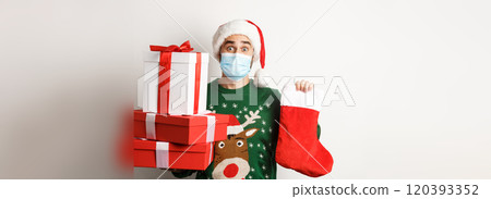 Winter holidays and xmas concept. Happy man in face mask and santa hat bringing gifts, holding Christmas sock and present boxes, standing over white background Winter holidays and xmas concept. Happy man in face mask and santa hat bringing gifts, holding Christmas sock and present boxes, standing over white background 120393352