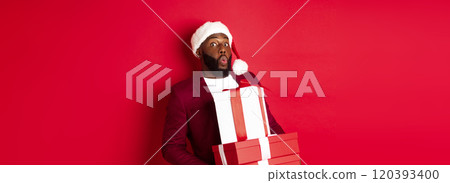 Christmas, New Year and shopping concept. Happy Black man in santa hat and blazer holding xmas presents, bring gifts and smiling, standing against red background 120393400