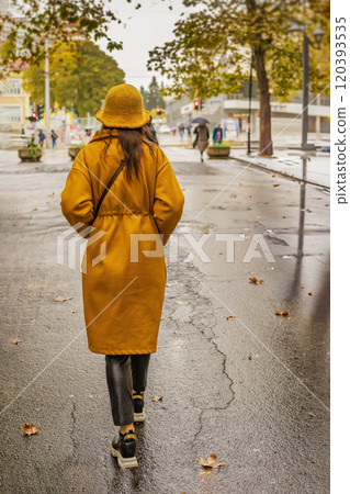 Woman in a yellow coat and hat walking on rainy street, rear view 120393535