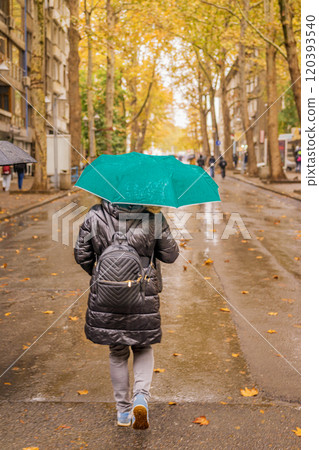 Back view of woman walking with green umbrella in an autumn street with fallen leaves 120393540