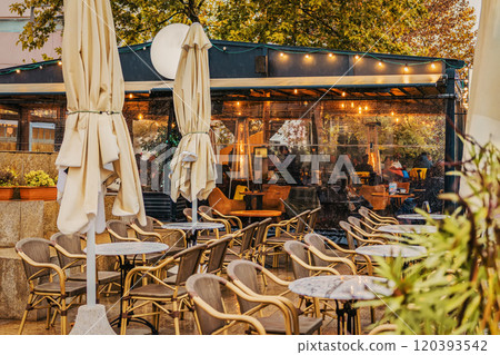 Outdoor cafe , closed umbrellas, warm lights glowing from the interior on rainy autumn evening Outdoor cafe , closed umbrellas, warm lights glowing from the interior on rainy autumn evening 120393542