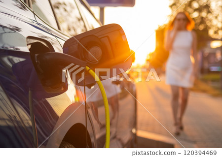 Woman in white dress walks by an electric car charging at sunset in a vibrant city street Woman in white dress walks by an electric car charging at sunset in a vibrant city street 120394469