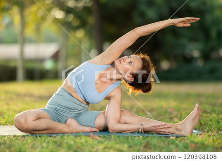 Woman practicing hatha yoga in garden, stretching in an asymmetrical seated asana Parivritta Janu Sirsasana Woman practicing hatha yoga in garden, stretching in an asymmetrical seated asana Parivritta Janu Sirsasana 120398108