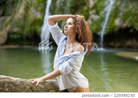 Brunette in lace panties and open shirt leaning against rock by waterfall 120398130