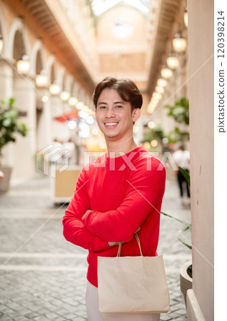 A handsome Asian man carrying a shopping bag, standing in a stunning shopping mall during winter. 120398214