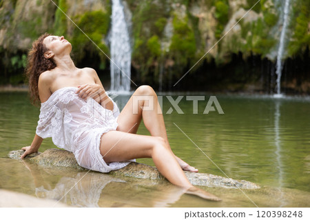 Half-naked girl in white translucent negligee poses sitting in nature against backdrop of waterfall 120398248