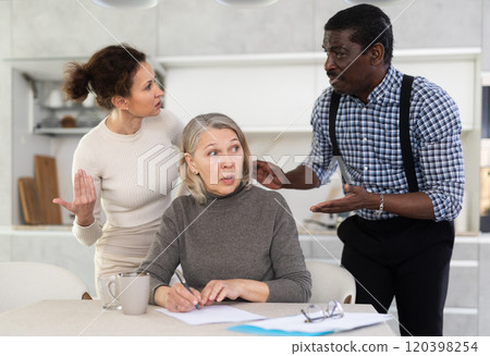 Old woman compiling testament while middle-aged man and woman competing with each other in the kitchen 120398254