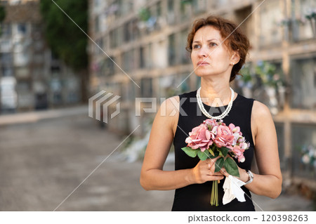 Sad woman in black dress with bouquet of flowers and handkerchief in hands, stands in cemetery 120398263