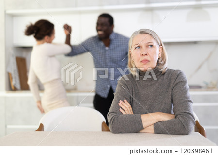 Old woman sitting at the kitchen table with her back to couple quarreling together Old woman sitting at the kitchen table with her back to couple quarreling together 120398461