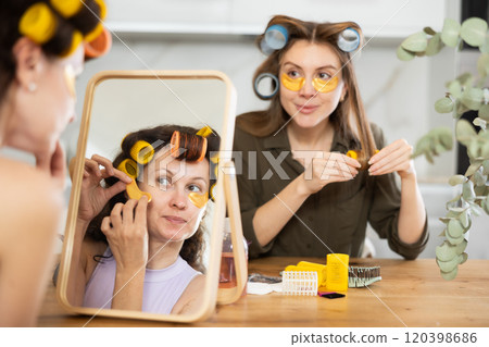 Woman in colorful curlers applying under eye patches by mirror with bestie 120398686
