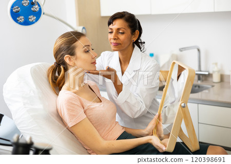 Young woman holding mirror during cosmetologist's examination Young woman holding mirror during cosmetologist's examination 120398691
