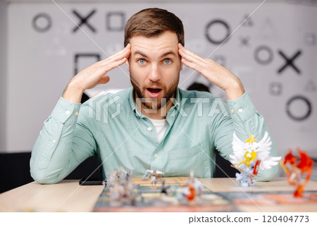 Man with board game piece while practicing strategy in home alone. A man plays a board game with cards and figures, expressing different emotions Man with board game piece while practicing strategy in home alone. A man plays a board game with cards and figures, expressing different emotions 120404773