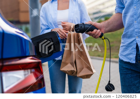 Man plugging in a connector to EV while his wife holds shopping bags at an urban parking lot. 120406825