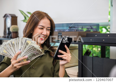 Employee Asian woman smiling she holding fan money dollar banknotes and using her mobile phone while sitting on a desk in home office, financial success and happiness 120407007