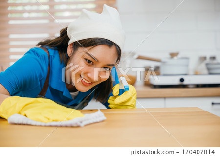 Asian cleaning service professional in yellow gloves wipes kitchen counter with liquid spray. Housekeeping assures home cleanliness and hygiene. Clean disinfect home care. maid household job. 120407014