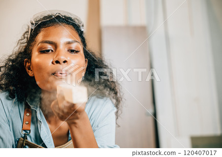 African American carpenter woman with curly hair and safety glasses blowing sawdust off wood in DIY workshop. Empowering female joiner confidently working in carpentry industry, National Carpenters African American carpenter woman with curly hair and safety glasses blowing sawdust off wood in DIY workshop. Empowering female joiner confidently working in carpentry industry, National Carpenters 120407017