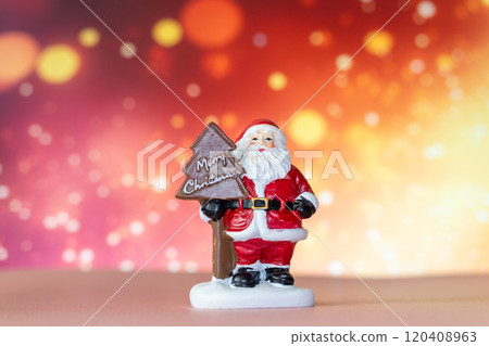 Santa Claus figurine holding a Merry Christmas tree sign, set against a glowing bokeh background in warm festive tones 120408963