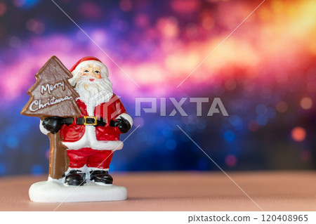 Santa Claus figurine holding a Merry Christmas tree sign, set against a glowing bokeh background in warm festive tones 120408965