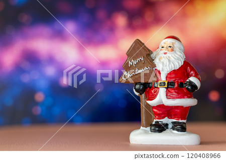 Santa Claus figurine holding a Merry Christmas tree sign, set against a glowing bokeh background in warm festive tones 120408966