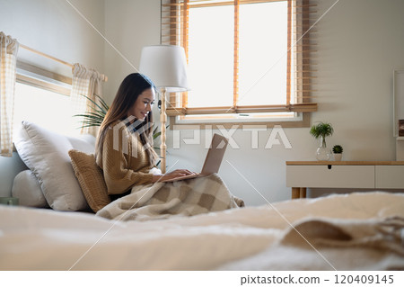 Beautiful young woman using laptop while sitting covered in blanket in bedroom Beautiful young woman using laptop while sitting covered in blanket in bedroom 120409145