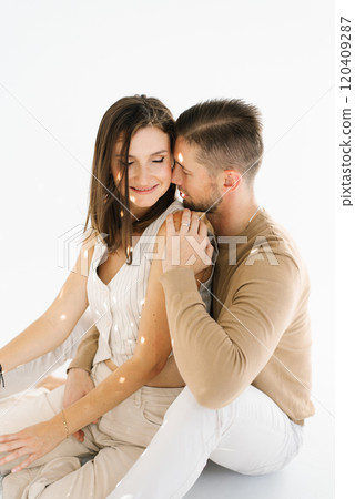 Young man and woman are sitting on a white surface, hugging each other. Scene is warm and affectionate 120409287