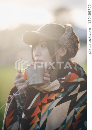 A woman drinking a drink while wrapped in a blanket at a campsite A woman drinking a drink while wrapped in a blanket at a campsite 120409703