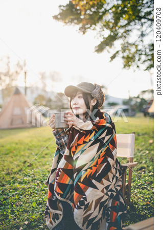 A woman drinking a drink while wrapped in a blanket at a campsite A woman drinking a drink while wrapped in a blanket at a campsite 120409708