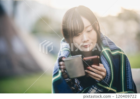A woman drinking a drink while wrapped in a blanket at a campsite 120409722