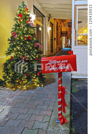 Red mailbox for Santa Claus letters near beautifully decorated Christmas tree with lights and bows. Lettere per Babbo Natale - Letters for Santa Claus 120416815