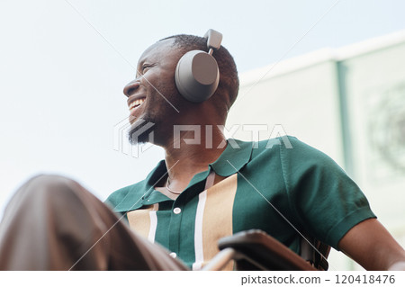 Candid low angle shot of young man with disability wearing headphones and listening to music in city setting smiling happily and looking away 120418476
