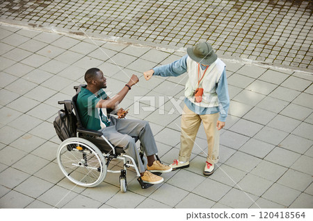Minimal high angle shot of African American man using wheelchair smiling happily and fist bumping with friend on graphic pavement tiles copy space 120418564