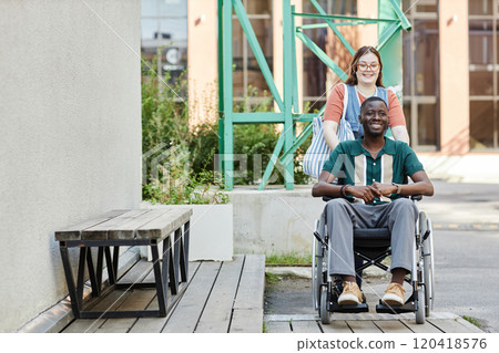 Front view portrait of smiling African American man with disability using wheelchair while moving up on ramp with friend or partner assisting copy space 120418576