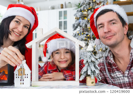Happy family in santa hats with tiny house in cozy home with Christmas decor of festive white kitchen. Gift for New Year. Insurance, moving to new house, mortgage, rent and purchase real estate Happy family in santa hats with tiny house in cozy home with Christmas decor of festive white kitchen. Gift for New Year. Insurance, moving to new house, mortgage, rent and purchase real estate 120419600