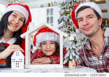 Happy family in santa hats with tiny house in cozy home with Christmas decor of festive white kitchen. Gift for New Year. Insurance, moving to new house, mortgage, rent and purchase real estate Happy family in santa hats with tiny house in cozy home with Christmas decor of festive white kitchen. Gift for New Year. Insurance, moving to new house, mortgage, rent and purchase real estate 120419601