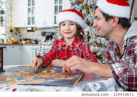 Happy family of dad and daughter in festive Santa hats Playing board game at table in a white kitchen with a Christmas tree and decor. New Year, family values. 120419651