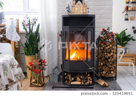 Metall black stove, burning hearth fireplace in white Festive interior of house is decorated for Christmas and New Year, Christmas tree. firewood in the woodpile, cozy and heating of home 120419691