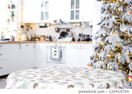 Round empty table to demonstrate the product with with tablecloth with pine needles and cones print, space for text in white kitchen, modern interior with a Christmas tree. New Year, Christmas Round empty table to demonstrate the product with with tablecloth with pine needles and cones print, space for text in white kitchen, modern interior with a Christmas tree. New Year, Christmas 120419704