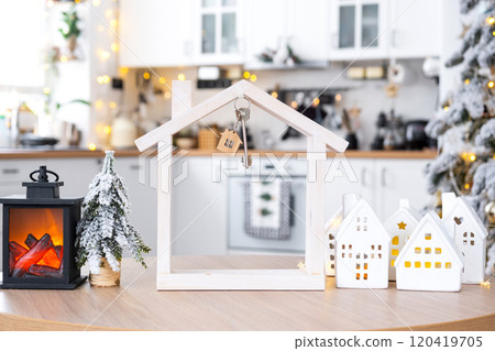 Key on Christmas tree and tiny house on cozy home with Christmas decor on table of festive white kitchen. Gift for New Year. Insurance, moving to new house, mortgage, rent and purchase real estate 120419705