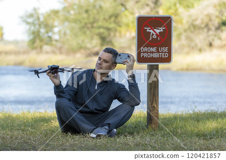 Operator is preparing to fly his quadcopter in national park no drone area. Man unlawfully using his UAV near restriction notice sign 120421857