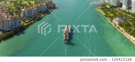 Commercial container ship entering Miami port harbor through main channel near South Beach. Luxurious hotels and residential buildings on waterfront and high skyscraper towers of downtown in distance Commercial container ship entering Miami port harbor through main channel near South Beach. Luxurious hotels and residential buildings on waterfront and high skyscraper towers of downtown in distance 120423188
