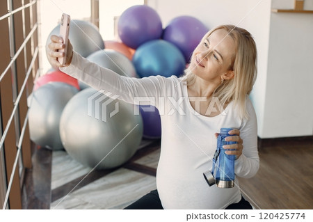 Caucasian pregnant woman in third trimester make a selfie and holding a bottle with water. Woman in bright fitness studio with big windows. Blonde woman wearing whit shirt and black leggins. Caucasian pregnant woman in third trimester make a selfie and holding a bottle with water. Woman in bright fitness studio with big windows. Blonde woman wearing whit shirt and black leggins. 120425774