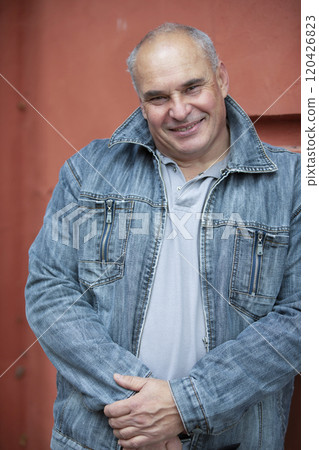 An elderly man in a denim jacket looks at the camera and smiles. An upright fifty-year-old gray-haired man of large build. 120426823